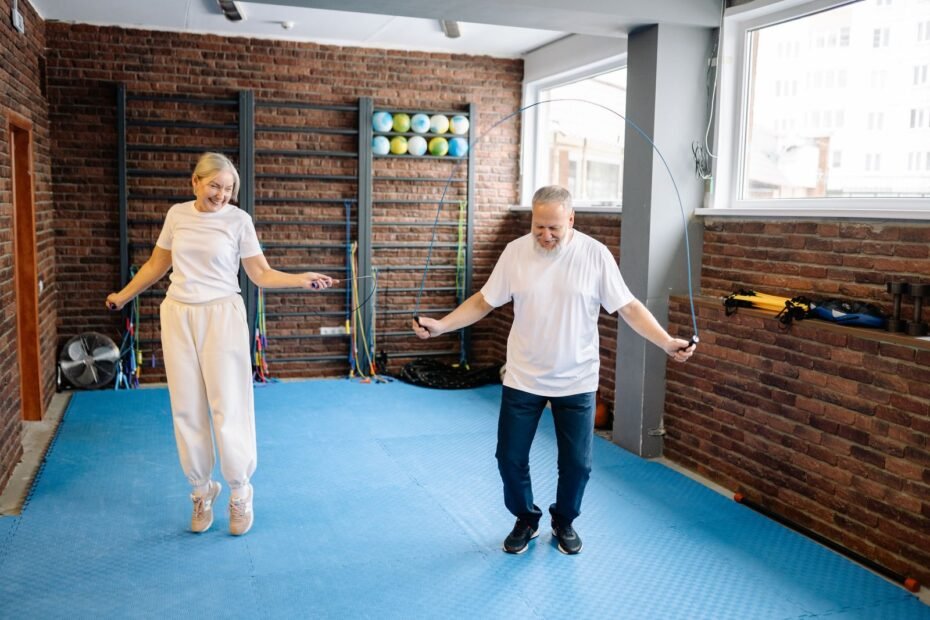 An older man and woman skipping with jump ropes on a padded gym floor.
