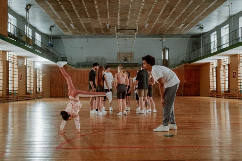 A coach watching closely as a young student practises a handstand-based movement during a group class in a sports hall.