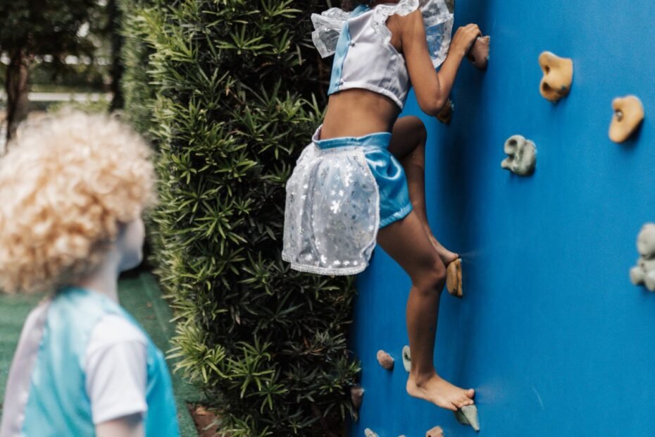 A child climbing a blue climbing wall on a playground while another child watches.