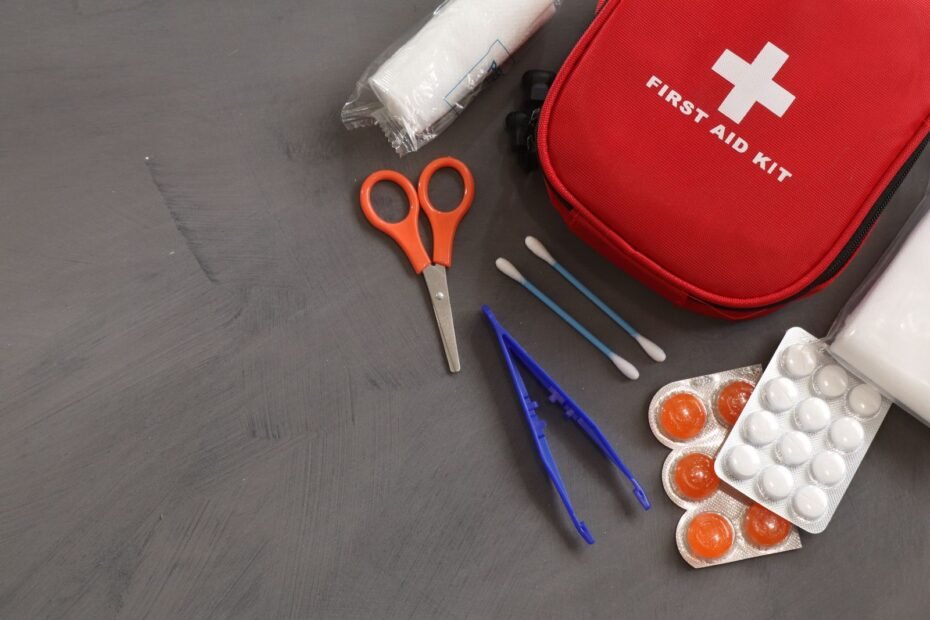 A red first aid kit with scissors, tweezers, cotton buds and a roll of bandage on a grey surface.
