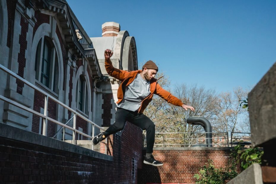 A traceur in an orange jacket dropping off a brick wall, arms out for balance before landing.