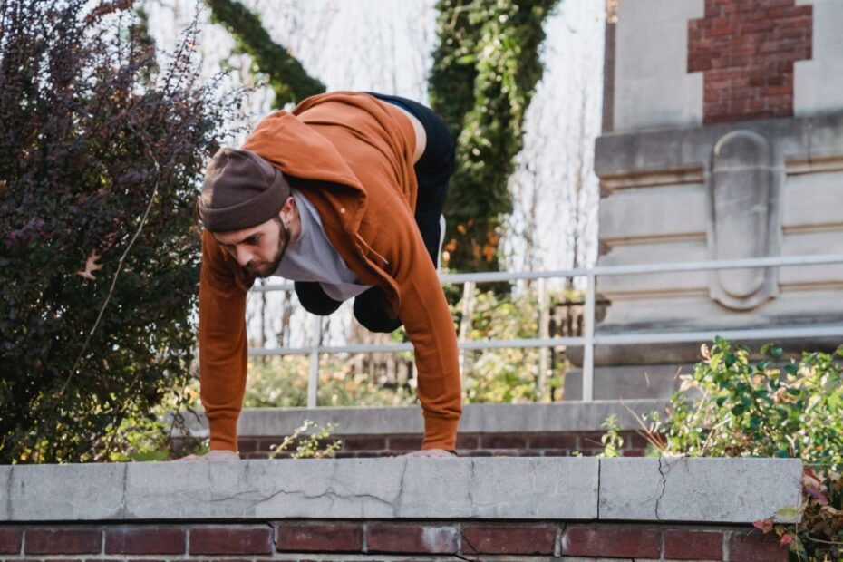 A traceur in an orange hoodie leaning forward with both hands planted flat on top of a low stone wall.
