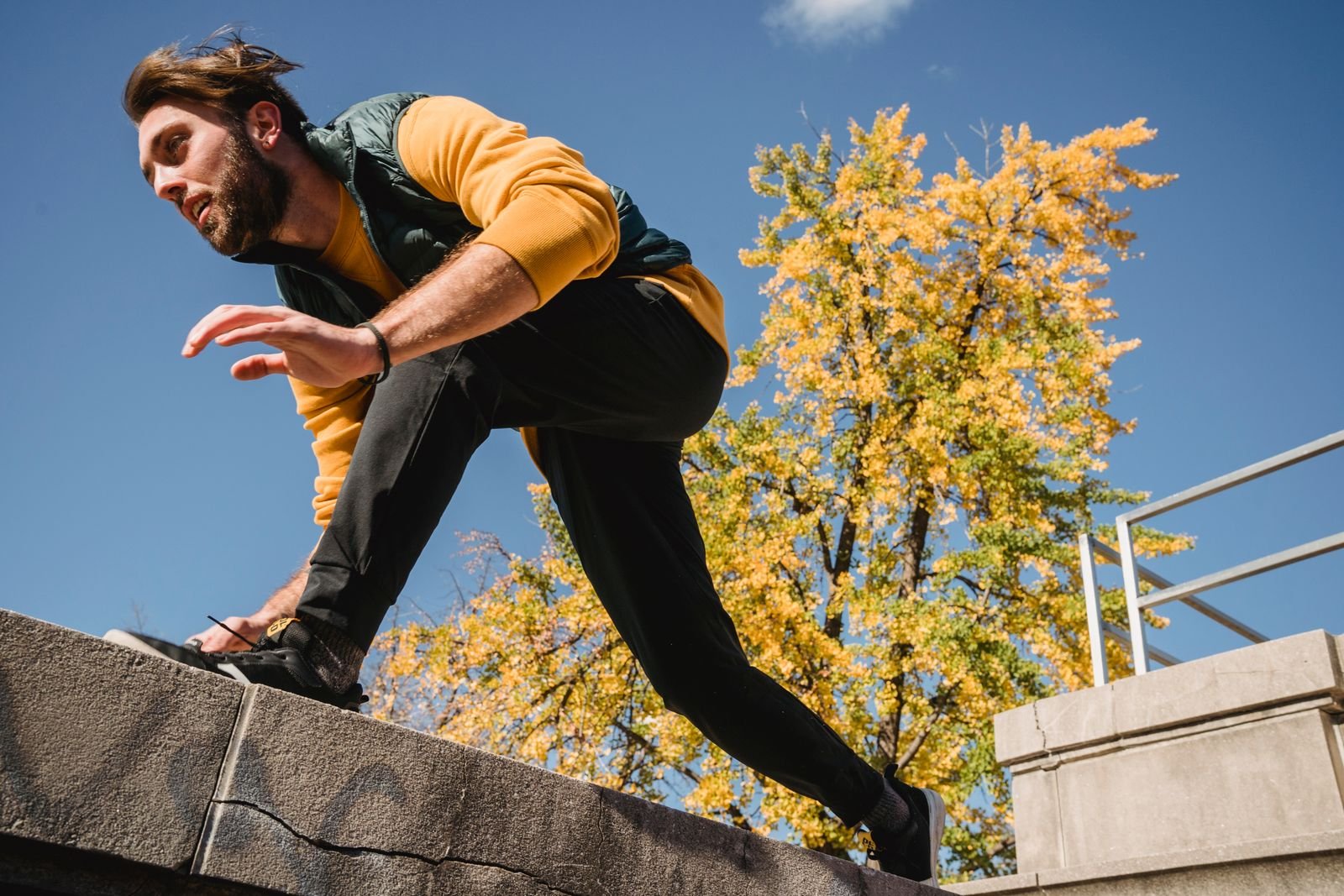 A traceur in a yellow top and green vest stepping over a stone wall, seen from below against a blue sky.