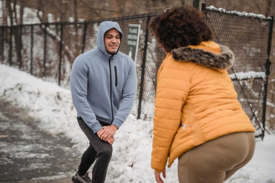 A man and a woman in warm layers doing a warm-up stretch outdoors next to snow.